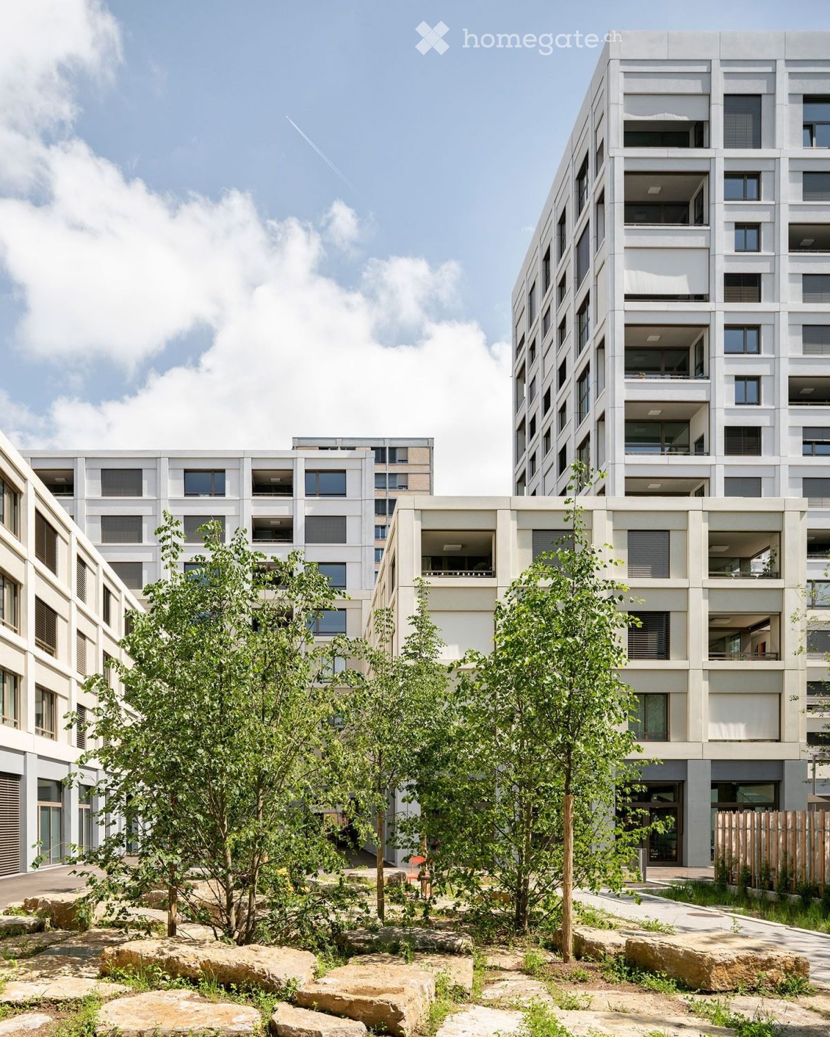 Modern multi-story residential building with balconies and large windows, surrounded by trees and stone pathways.