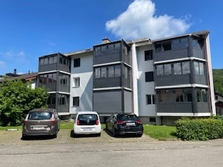 Modern multi-story apartment building with large windows and balconies, three cars parked in front.