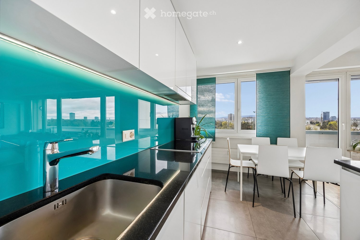Modern kitchen with a turquoise backsplash, stainless steel sink, and a small dining area with white chairs and table.