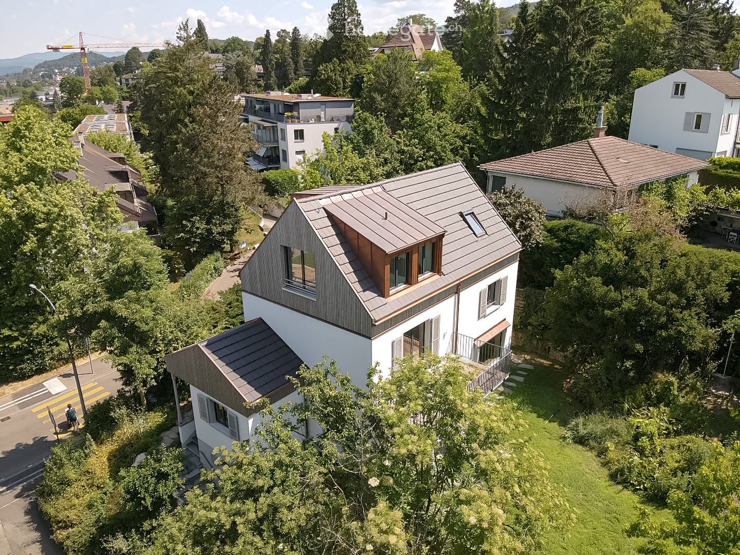 A modern two-story house with a grey metal roof and white walls, surrounded by lush greenery and trees. The house features multiple windows and a small balcony on the upper level.