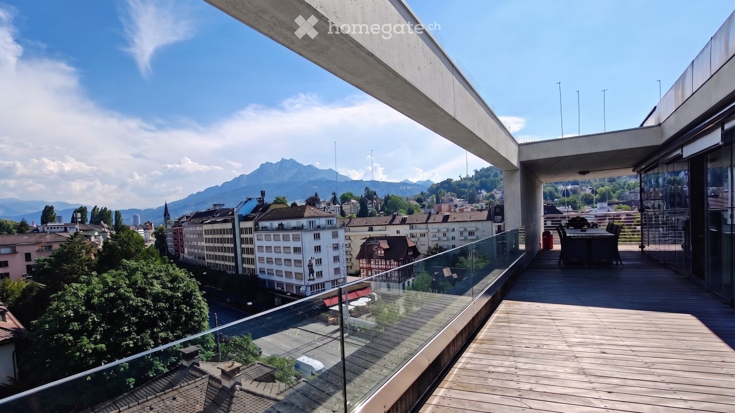 Balcony with wooden decking, glass railing, and outdoor furniture overlooking a cityscape with mountains in the background.