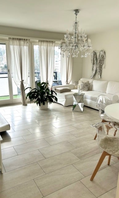 Modern living room with large windows, white sofa, coffee table, chandelier, and decorative plant.