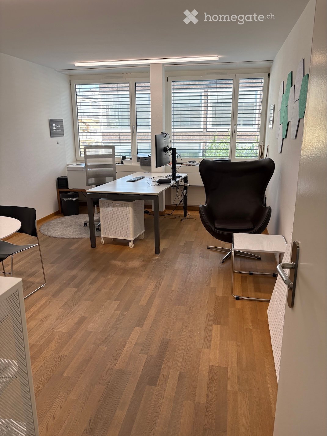 Office space with wooden flooring, large windows with blinds, a desk with a screen, chairs, and a small table.