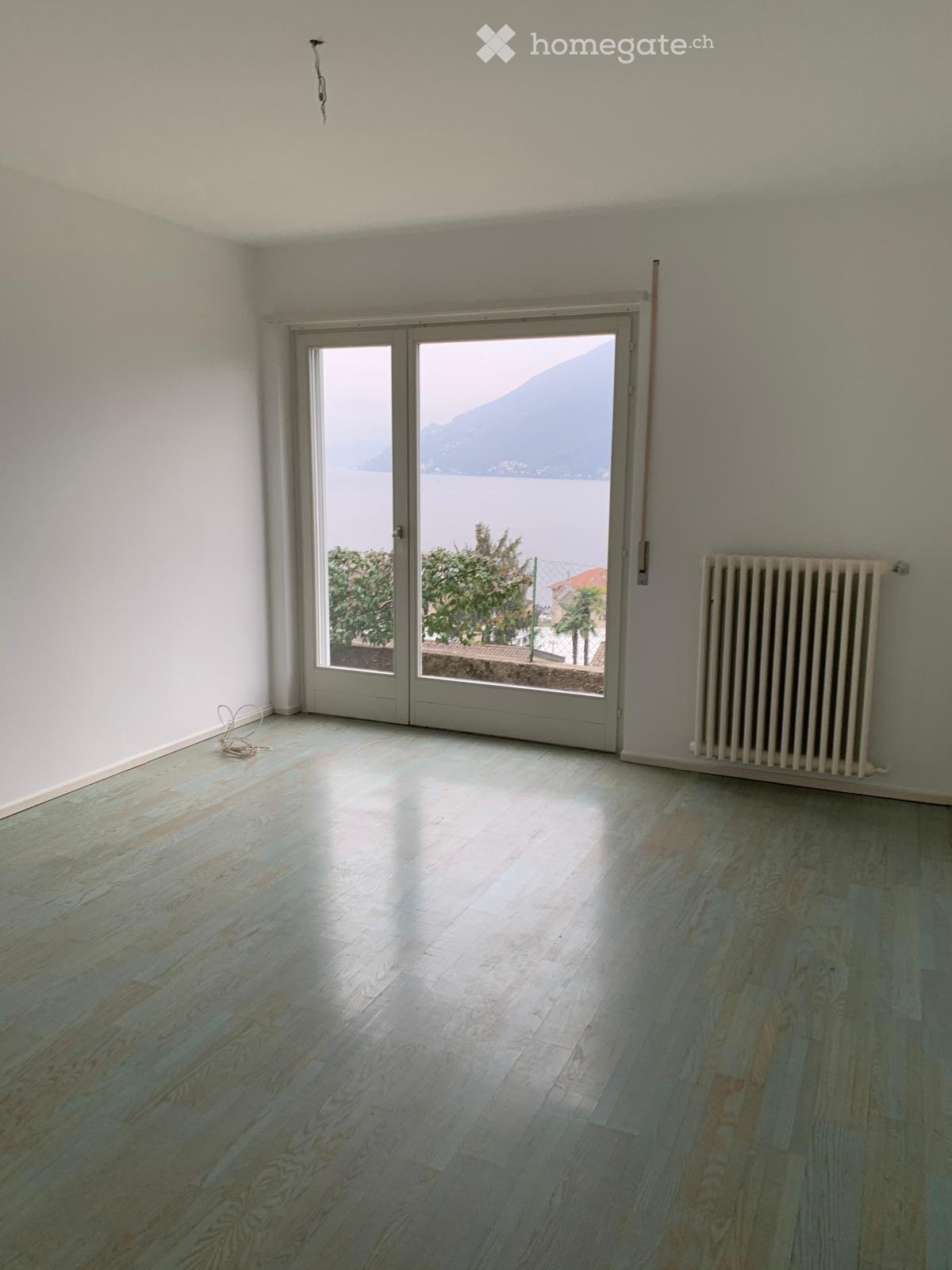 Empty room with light wooden flooring, white walls, large sliding glass doors leading to a scenic view of a lake and mountains, and a radiator on one wall.