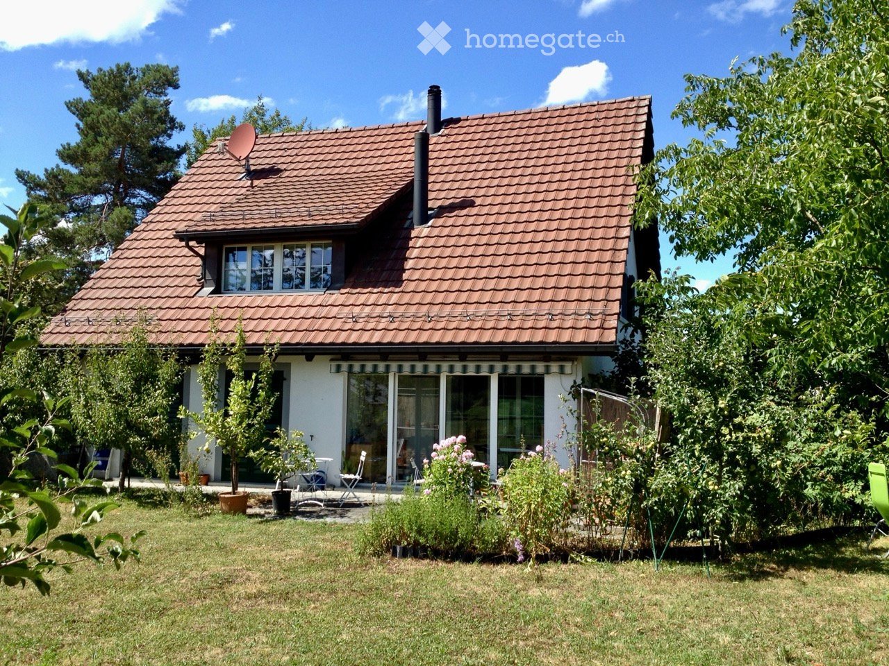 Two-story house with red-tiled roof, white walls, large windows, and a satellite dish on the roof. The house is surrounded by a well-maintained garden with trees, shrubs, and outdoor seating.