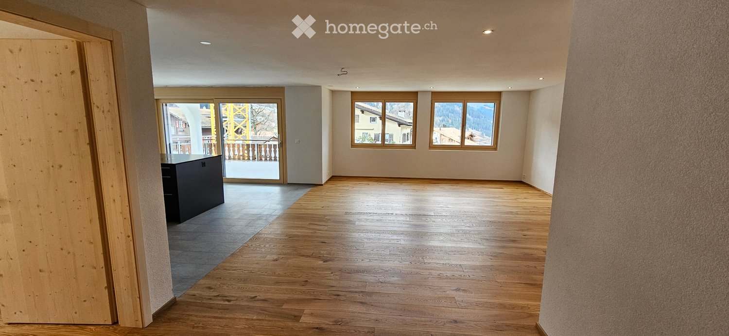 Empty room with wooden flooring, large windows, and a small black cabinet near the entrance. The space is well-lit with recessed ceiling lights.