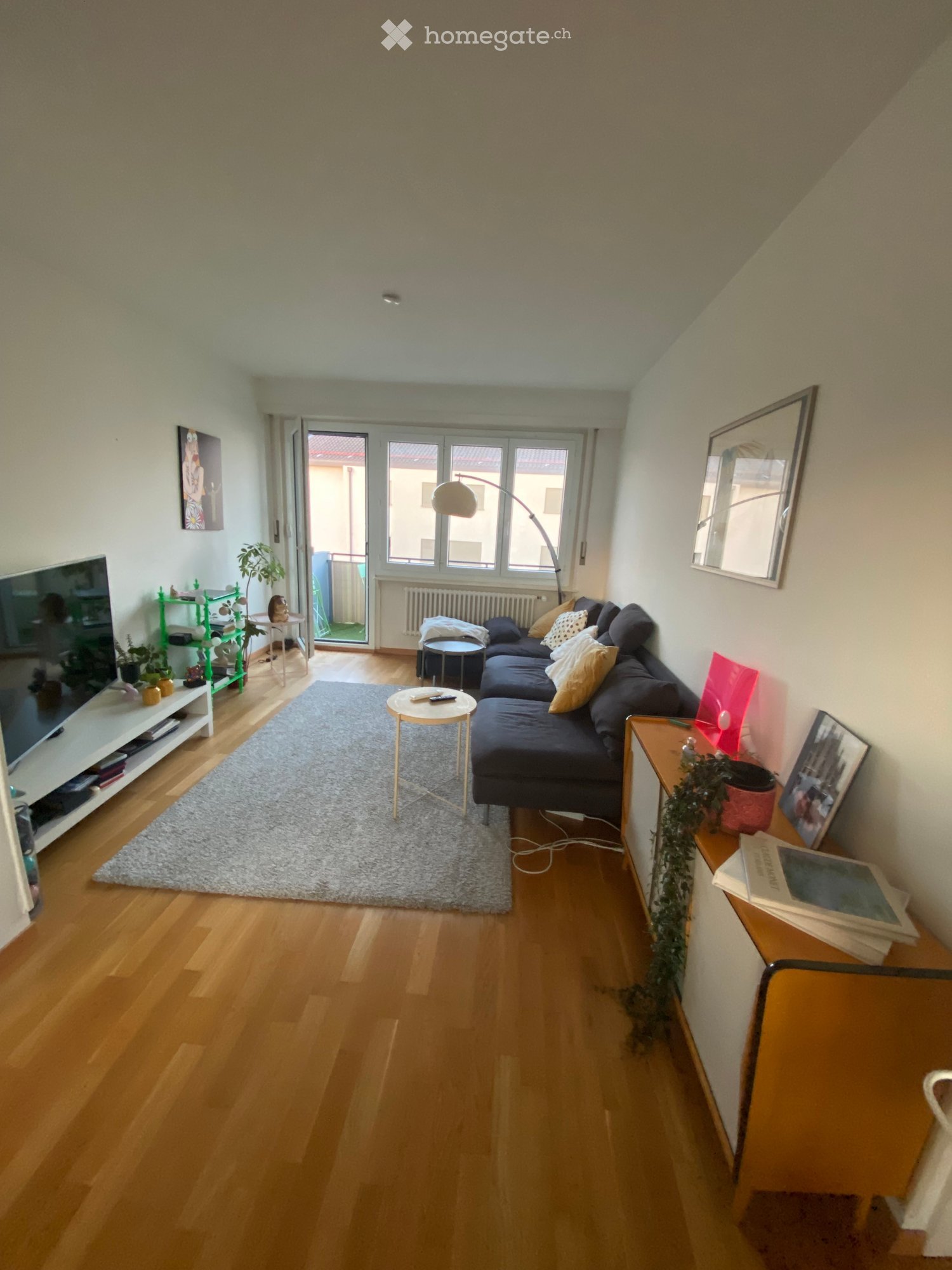 Modern living room with wooden flooring, grey rug, dark blue sofa, coffee table, side table, floor lamp, TV stand, and decorative items.