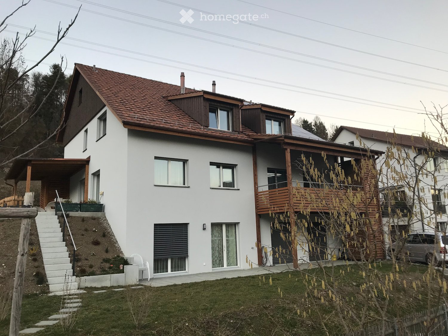 Two-story house with white exterior, red-tiled roof, multiple windows, raised ground floor, and balcony on the upper level.