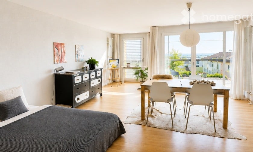 Bright, modern room with a bed, dresser, dining table, chairs, and a hanging lamp. Large windows offer a view of greenery outside.