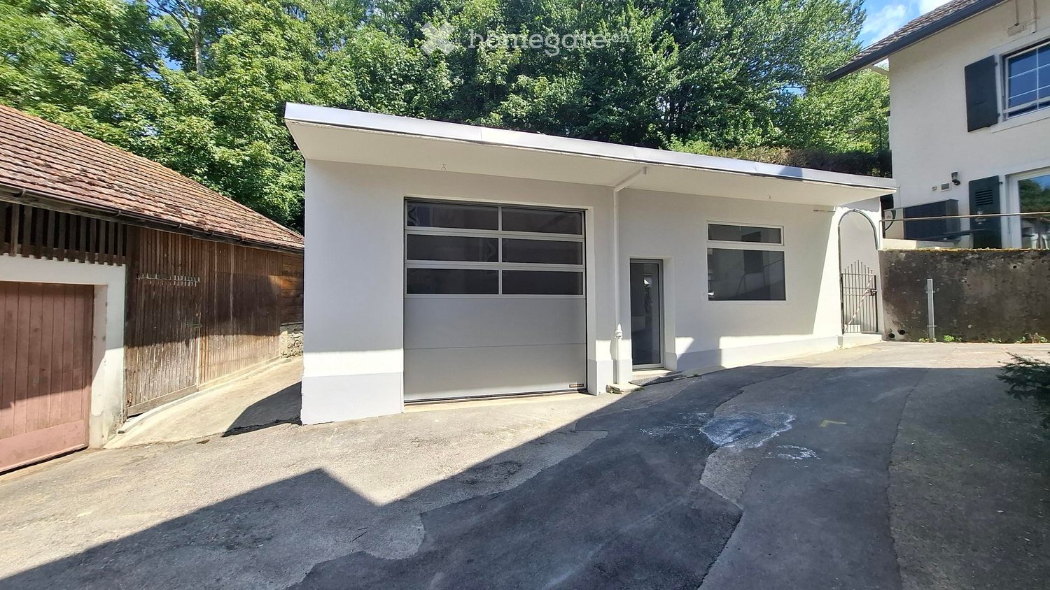 A modern white building with a large garage door and a separate wooden garage on the left side.