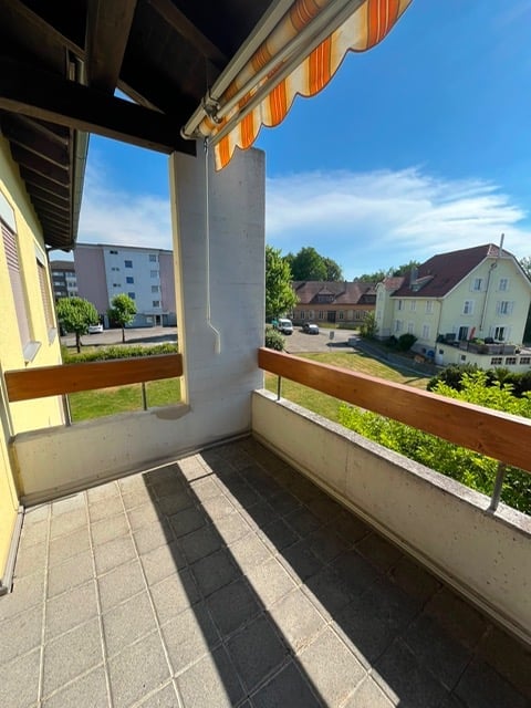 Balcony with tiled floor, wooden railings, striped awning, overlooking a residential area with houses and greenery.