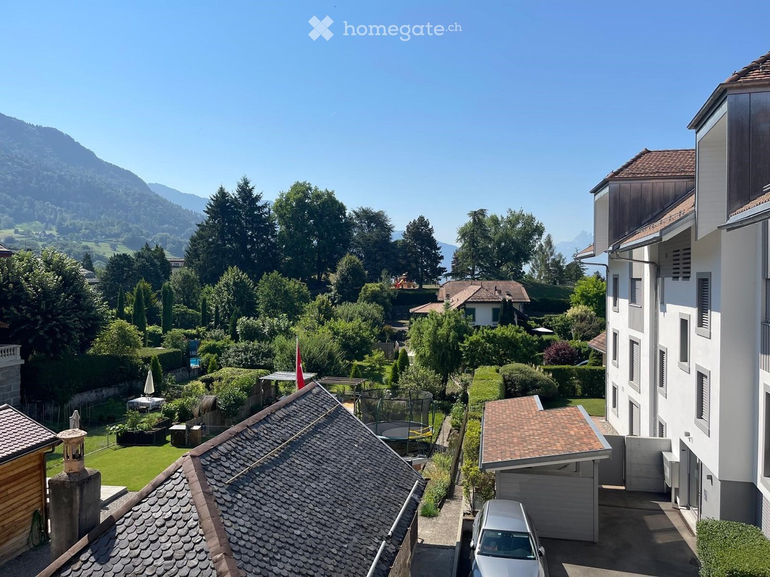 A peaceful garden area with a trampoline, a small table, and a Swiss flag, surrounded by trees and mountains.