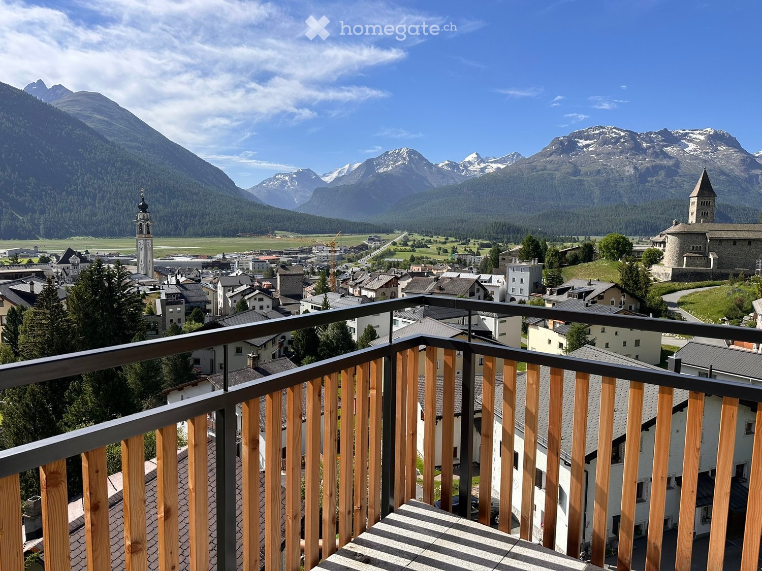 Balcony overlooking a picturesque town with traditional houses, a church tower, and snow-capped mountains in the background.