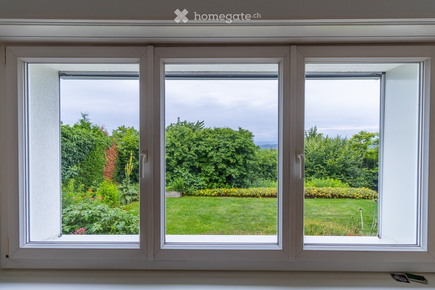 Three large windows with white frames open to a lush garden with green grass, bushes, and trees.