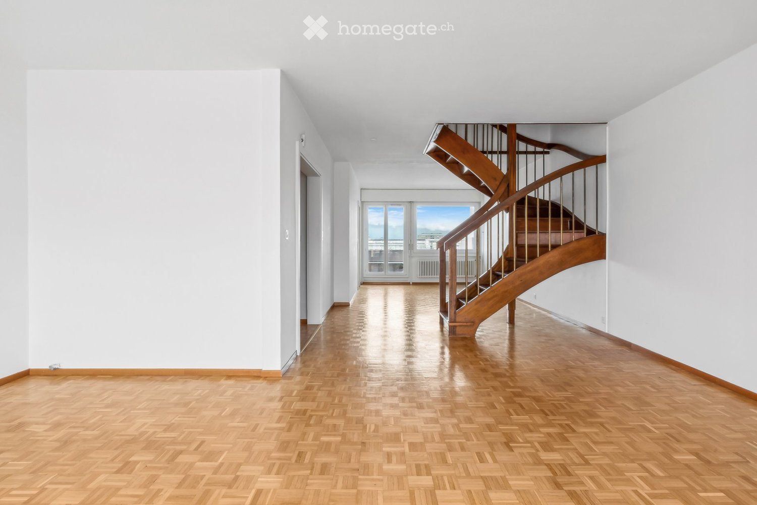Empty hallway with wooden flooring, white walls, and a wooden staircase leading upwards.