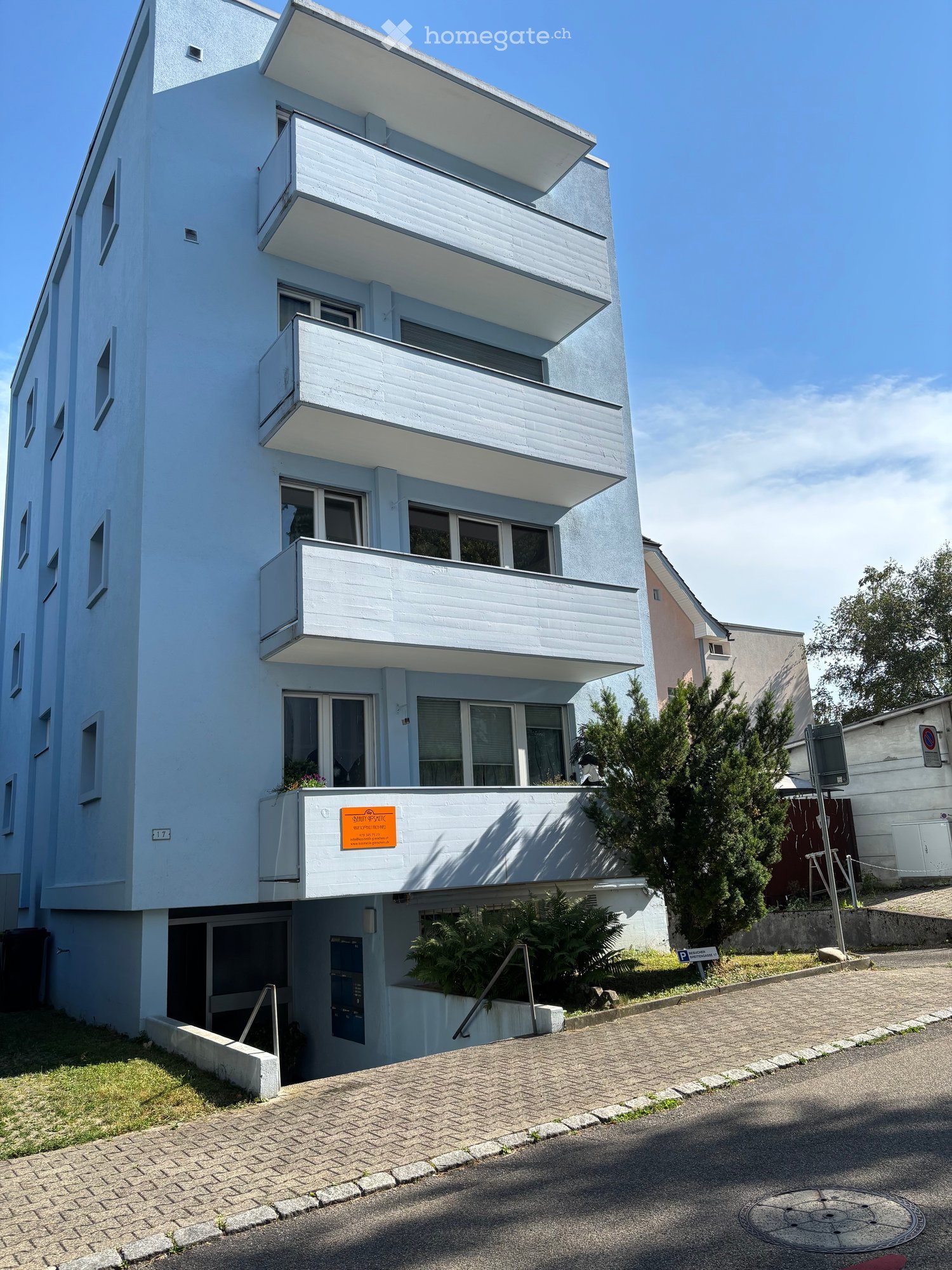 Modern multi-story building with light blue facade, white balconies, and large windows. Features a raised ground floor with greenery and a paved driveway.