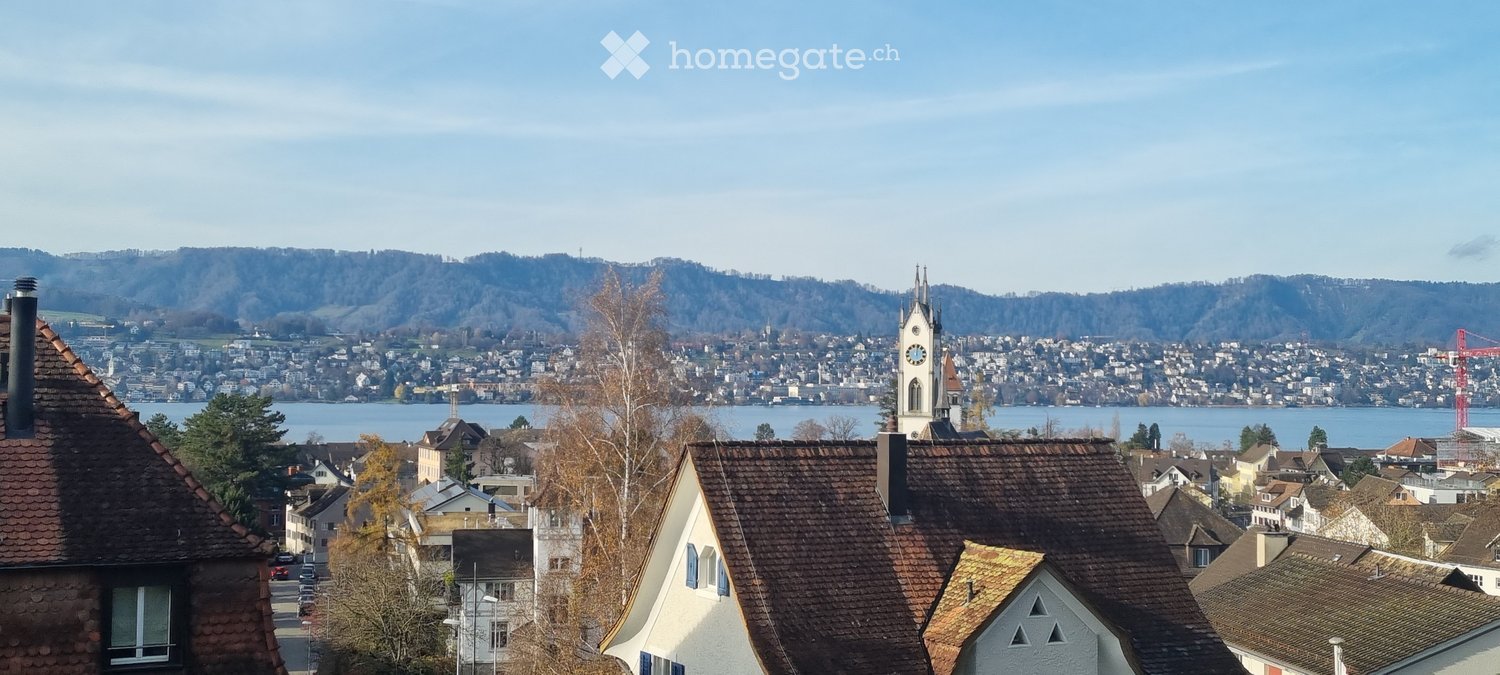 A panoramic view of a town with numerous houses, a church tower, and a lake in the background, surrounded by hills.