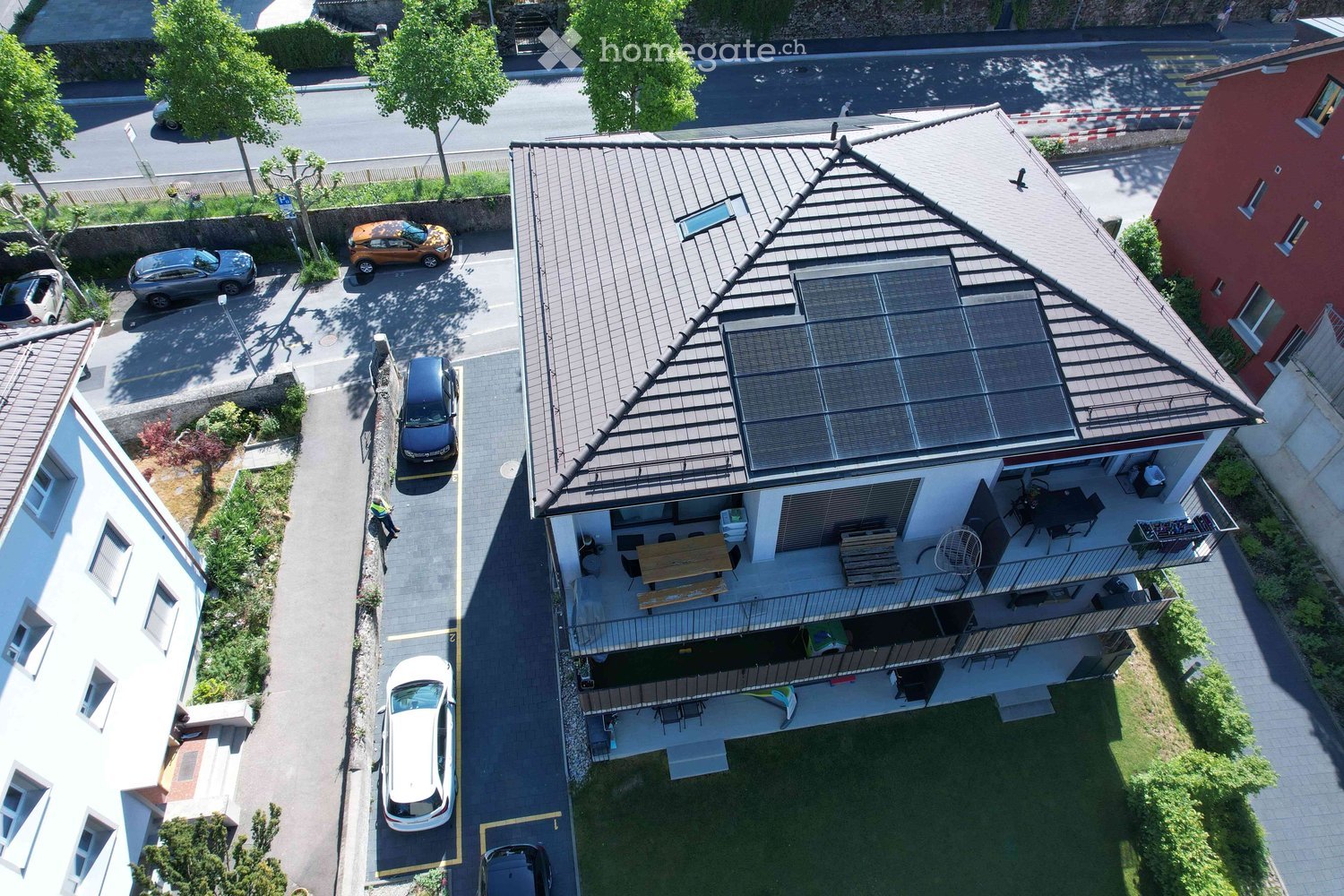 Aerial view of a modern house with a flat roof, solar panels, and a spacious balcony area with outdoor furniture. The house is surrounded by a parking area with several cars and greenery.