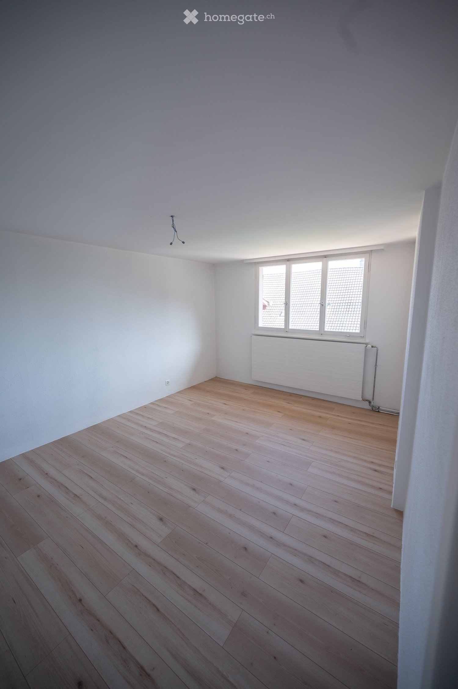 Empty room with light wooden flooring, white walls, and a window with blinds. A radiator is visible below the window.