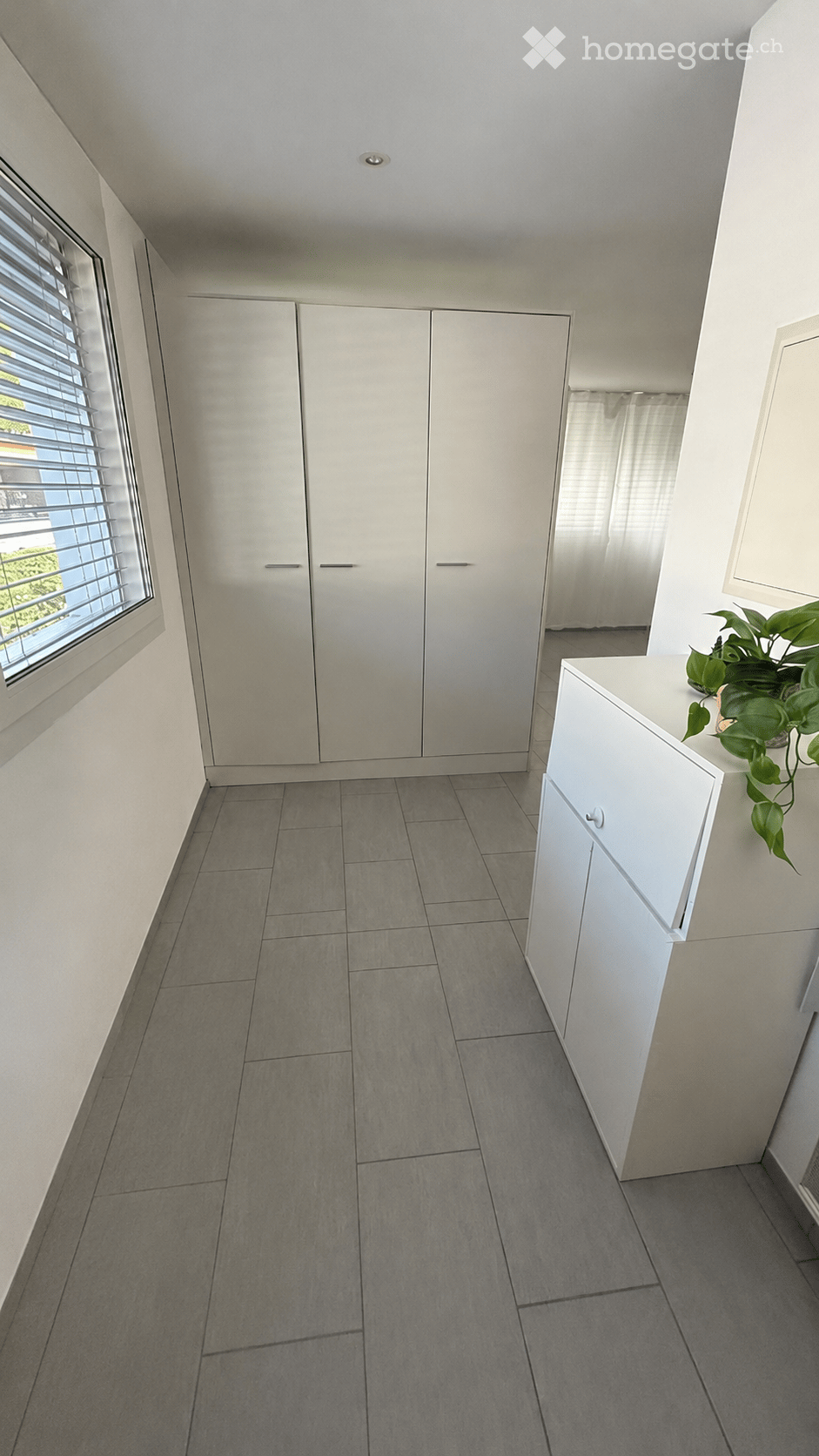 Empty room with grey tiled floor, white built-in wardrobes, a small white cabinet, and a window with blinds. A potted plant is visible on the cabinet.