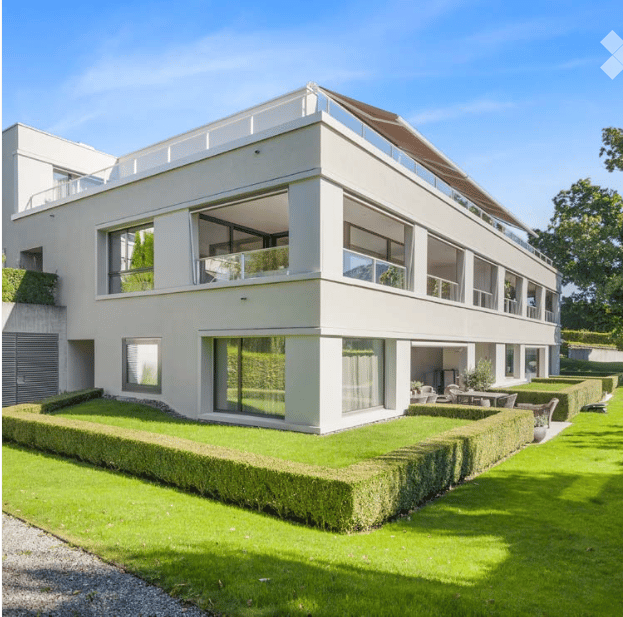 Modern two-story house with white exterior, large windows, and a flat roof. The house features a well-maintained lawn and neatly trimmed hedges in the front yard.