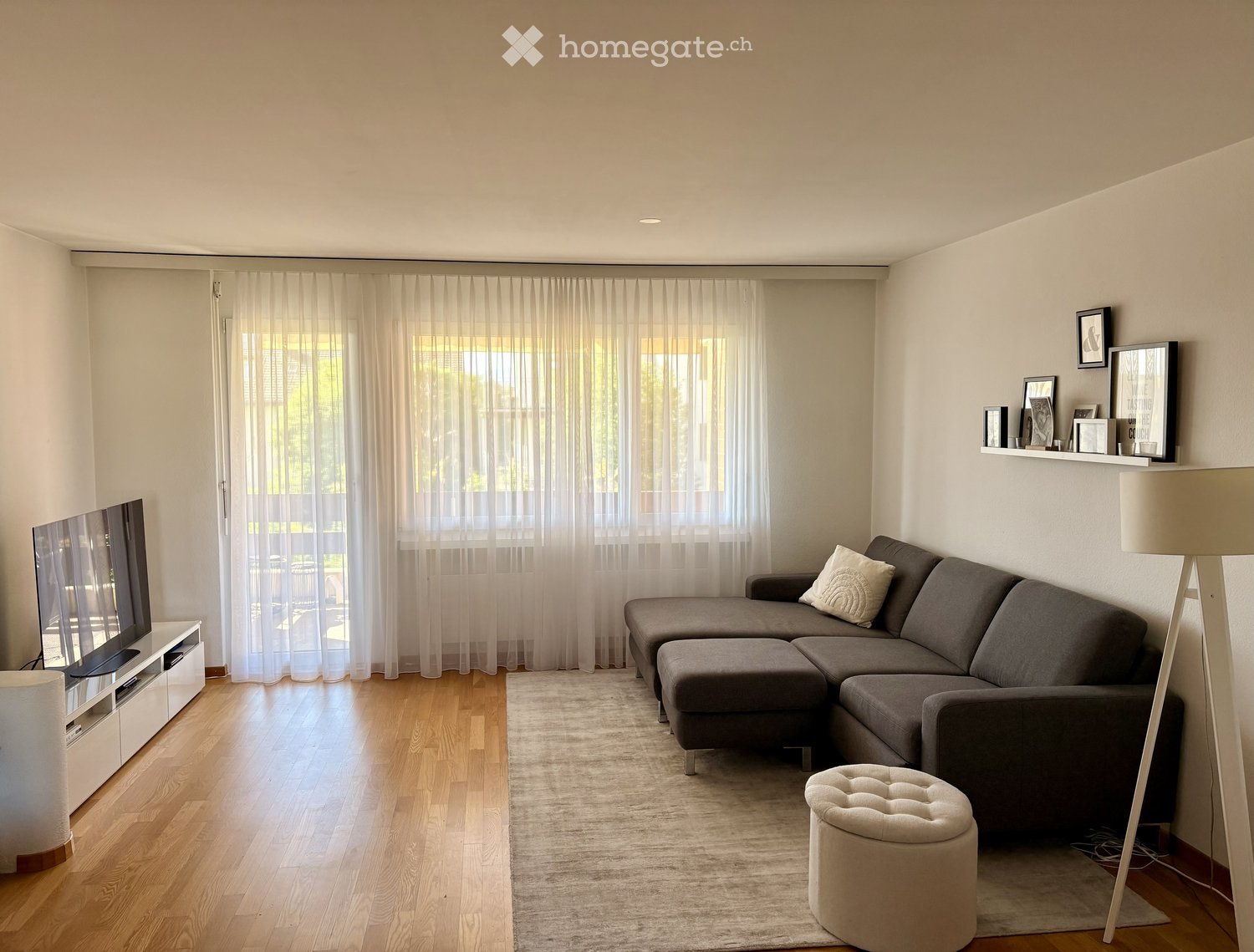 Modern living room with a grey sectional sofa, ottoman, cream-colored stool, television on a white stand, floor lamp, and a shelf with framed pictures.