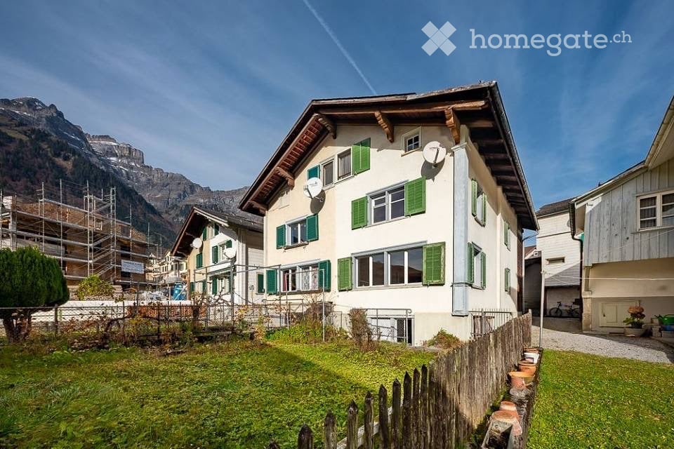 A two-story house with white walls and green shutters, surrounded by a small garden and a wooden fence, with a mountain view in the background.
