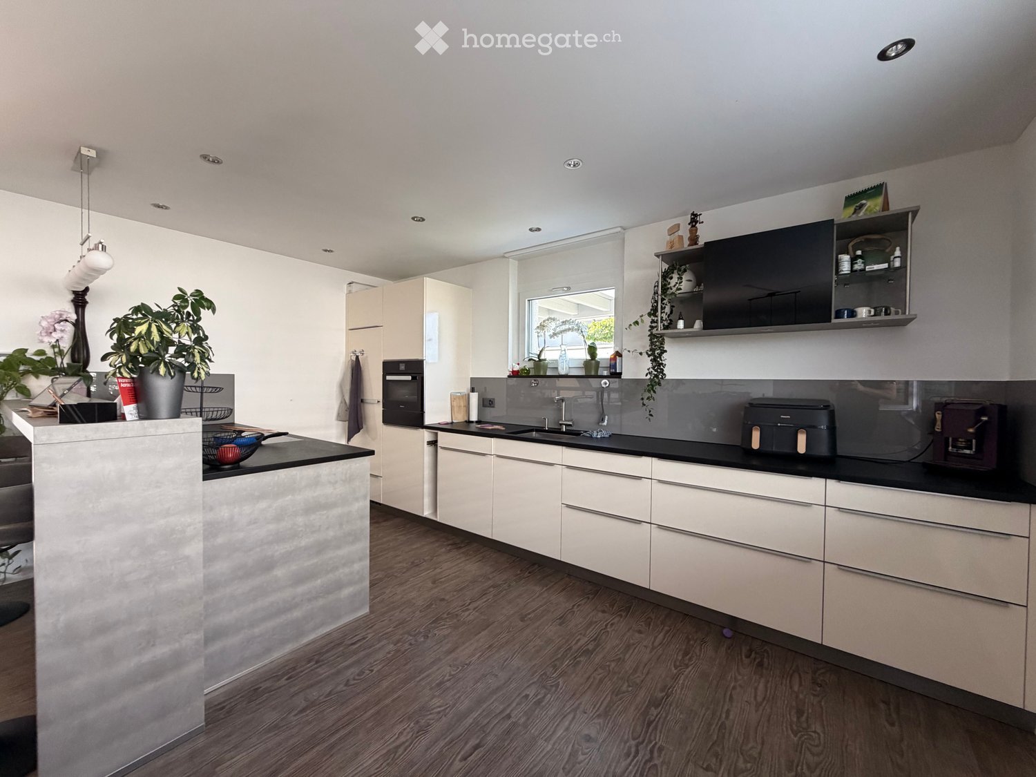 Modern kitchen with white cabinets, black countertops, and wooden flooring. Features include a sink, oven, printer, coffee maker, and decorative plants.