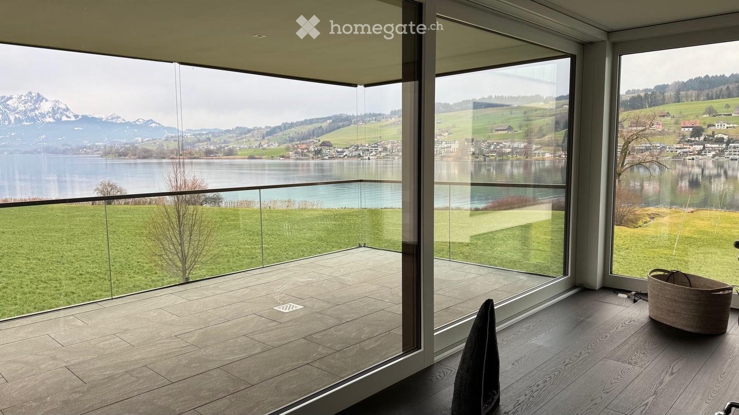 Modern balcony with large glass walls offering scenic views of a lake and mountains, featuring a wicker basket and a folded garment on the floor.