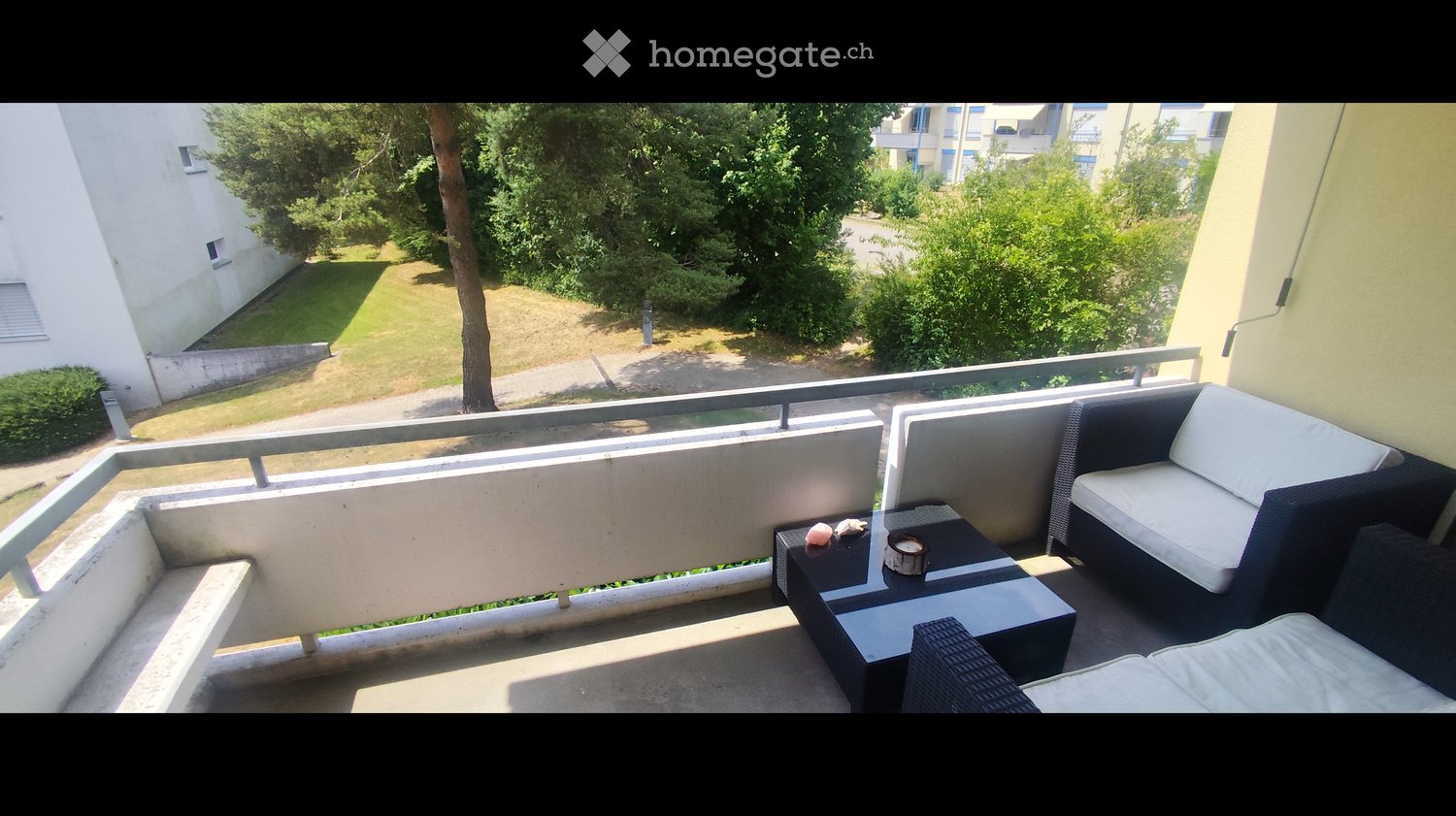 Balcony with a view of a green area, featuring a black sofa, a small table, and a wicker chair.