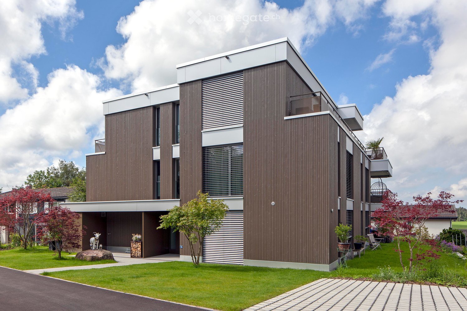 Modern two-story house with wooden siding and white trim, featuring large windows, a balcony, and a well-maintained garden with trees and shrubs.
