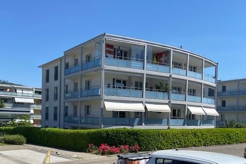 Modern multi-story apartment building with balconies, white awnings, and a neatly trimmed hedge in front.
