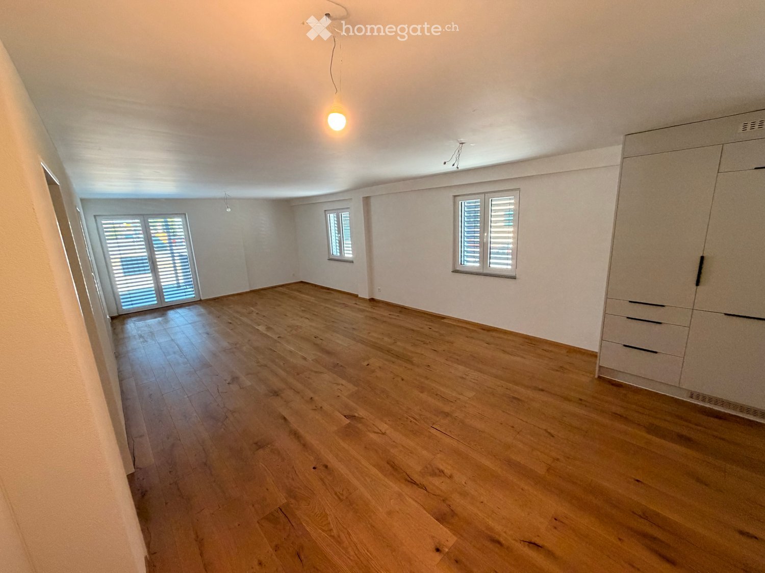 Empty room with wooden flooring, white walls, and built-in closet. Three windows with shutters provide natural light.