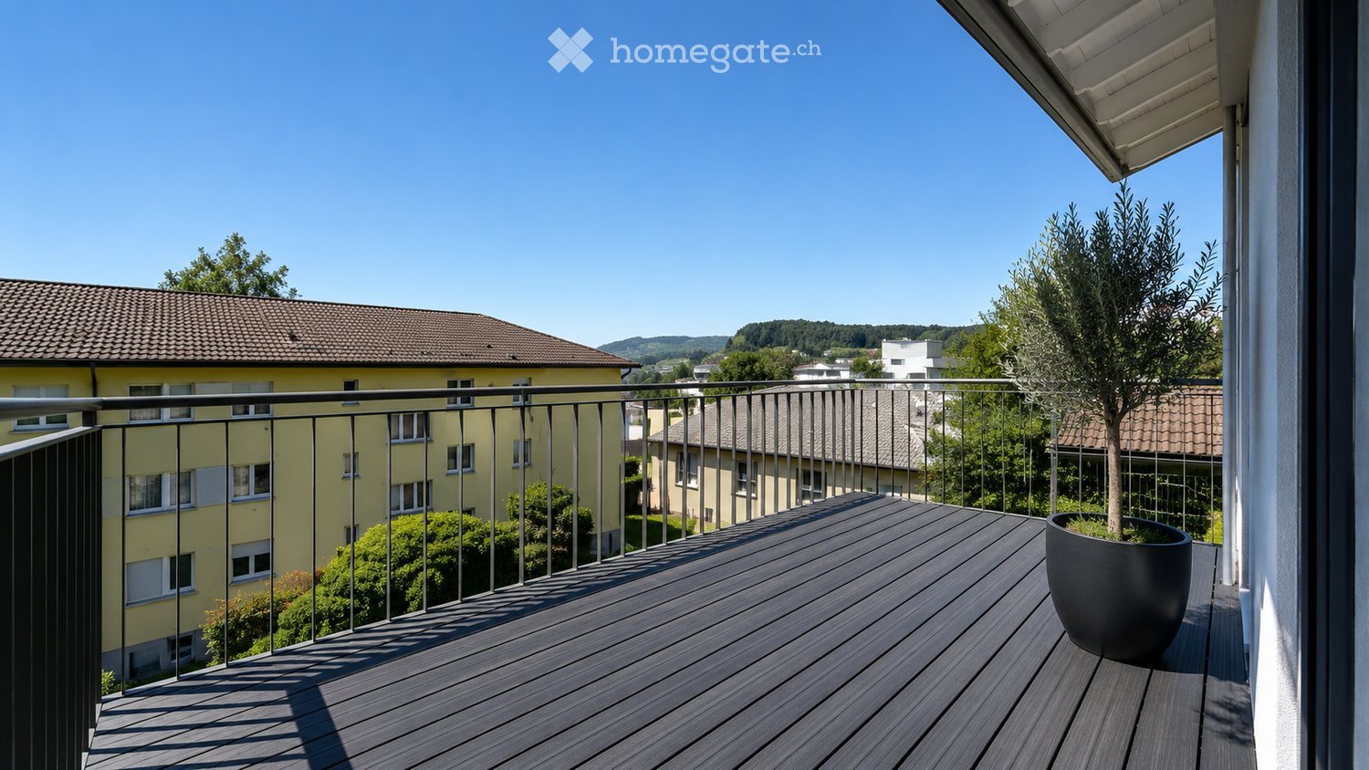 Balcony with wooden decking, metal railing, and a potted plant, overlooking a residential area with houses and trees.