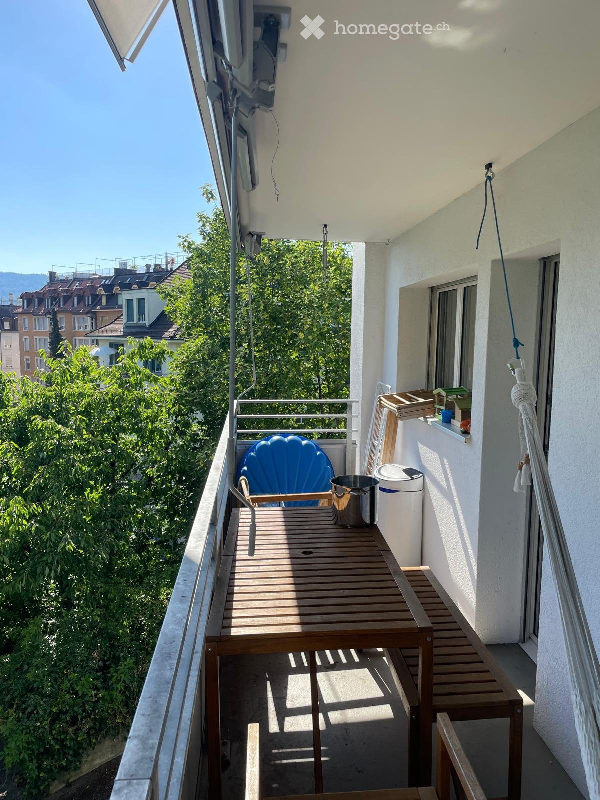 Balcony with wooden table, blue chair, potted plant, and railing overlooking greenery.