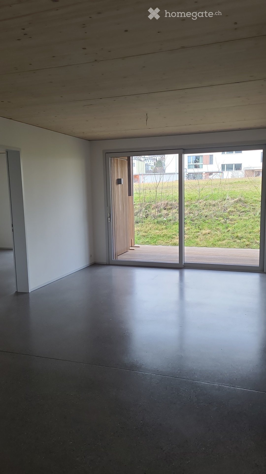 Empty room with concrete floor, white walls, and large sliding glass doors leading to a wooden deck and grassy area.
