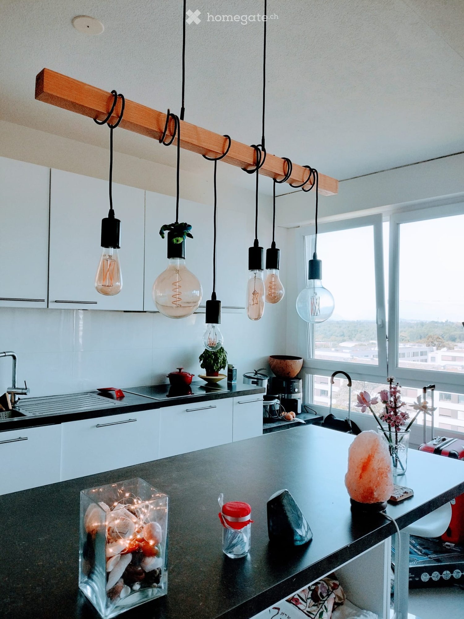 Modern kitchen with white cabinets, black countertops, and a large window offering a view. Features include a sink with a modern faucet, a coffee maker, a decorative bowl, a teapot, a flower vase, a salt lamp, a crystal, a glass container with lights, and