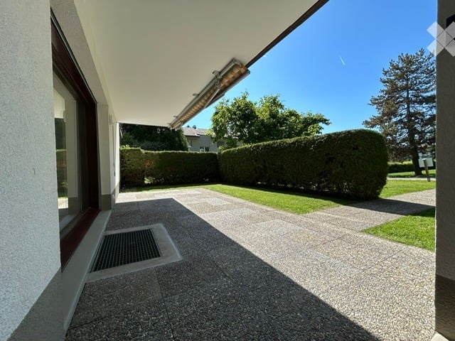 A modern outdoor patio area with a clean, paved surface, surrounded by neatly trimmed hedges and trees. The area is adjacent to a building with large windows and a covered walkway.
