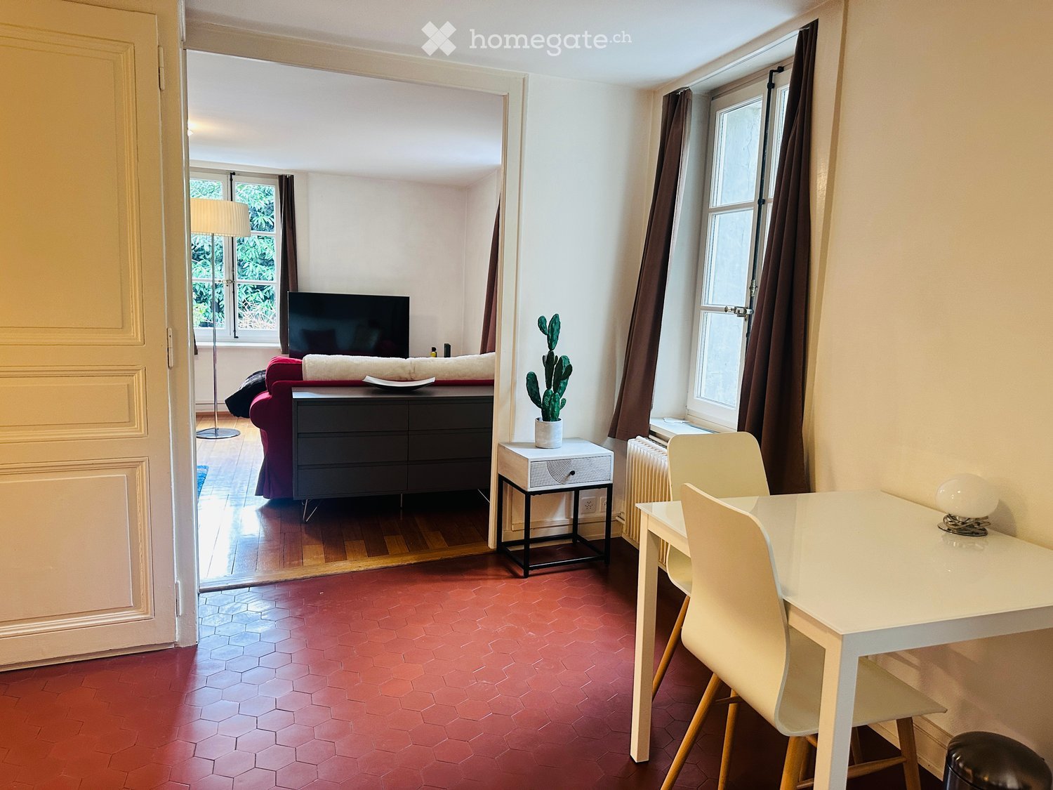A modern, well-lit living space with a red hexagonal tile floor, a white dining table with chairs, a sofa, a television, and a small side table with a plant.