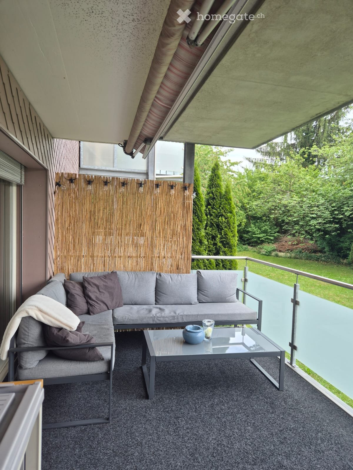 Balcony with grey sectional sofa, coffee table, decorative vase, and cup. Bamboo privacy screen, glass railing, and outdoor carpet.