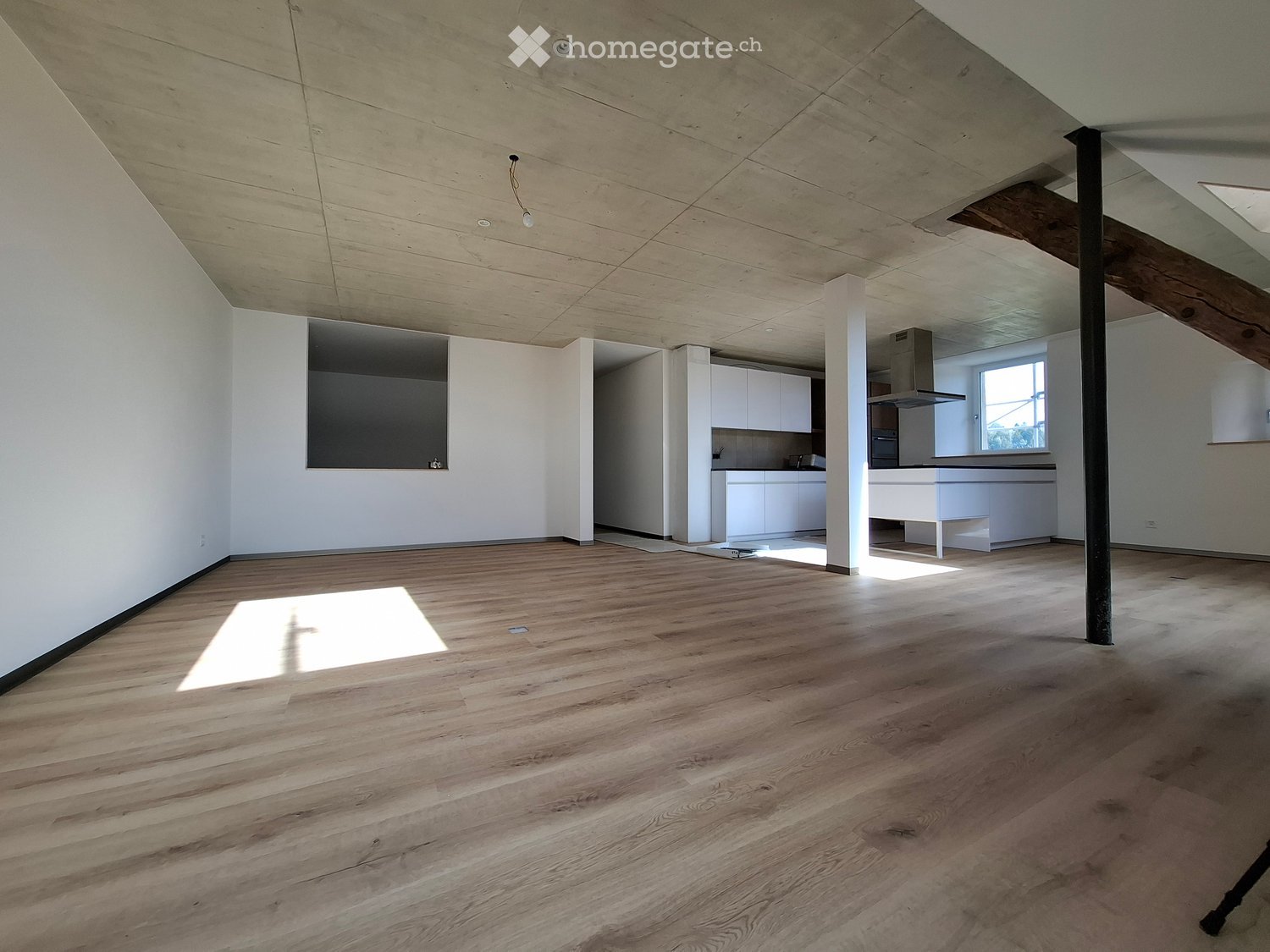 Open, unfurnished room with wooden flooring, white walls, and exposed wooden beams on the ceiling. The room features a large window allowing natural light to enter, and a small kitchen area is visible in the background.