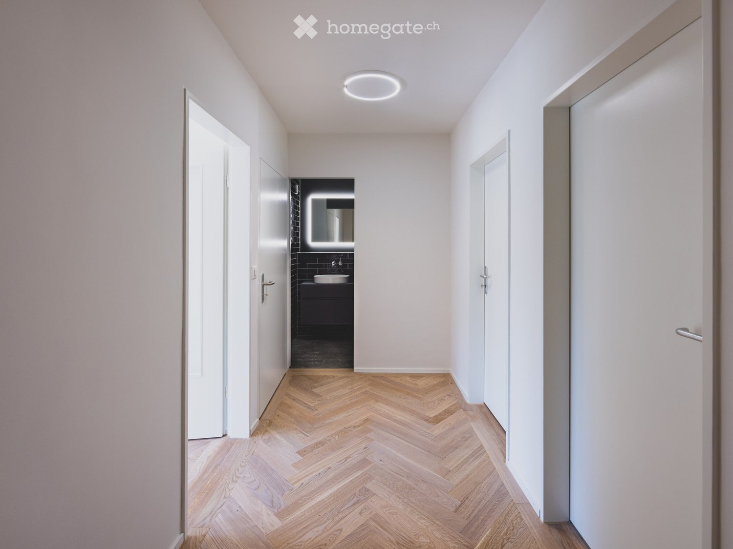 Modern hallway with herringbone wooden flooring, white walls, recessed ceiling light, and doors leading to rooms including a bathroom with dark tiles and a sink.