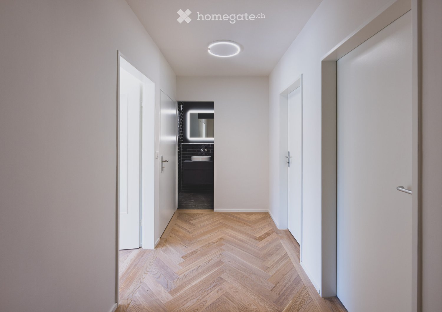 Modern hallway with herringbone wooden flooring, white walls, recessed ceiling light, and doors leading to rooms including a bathroom with dark tiles and a sink.