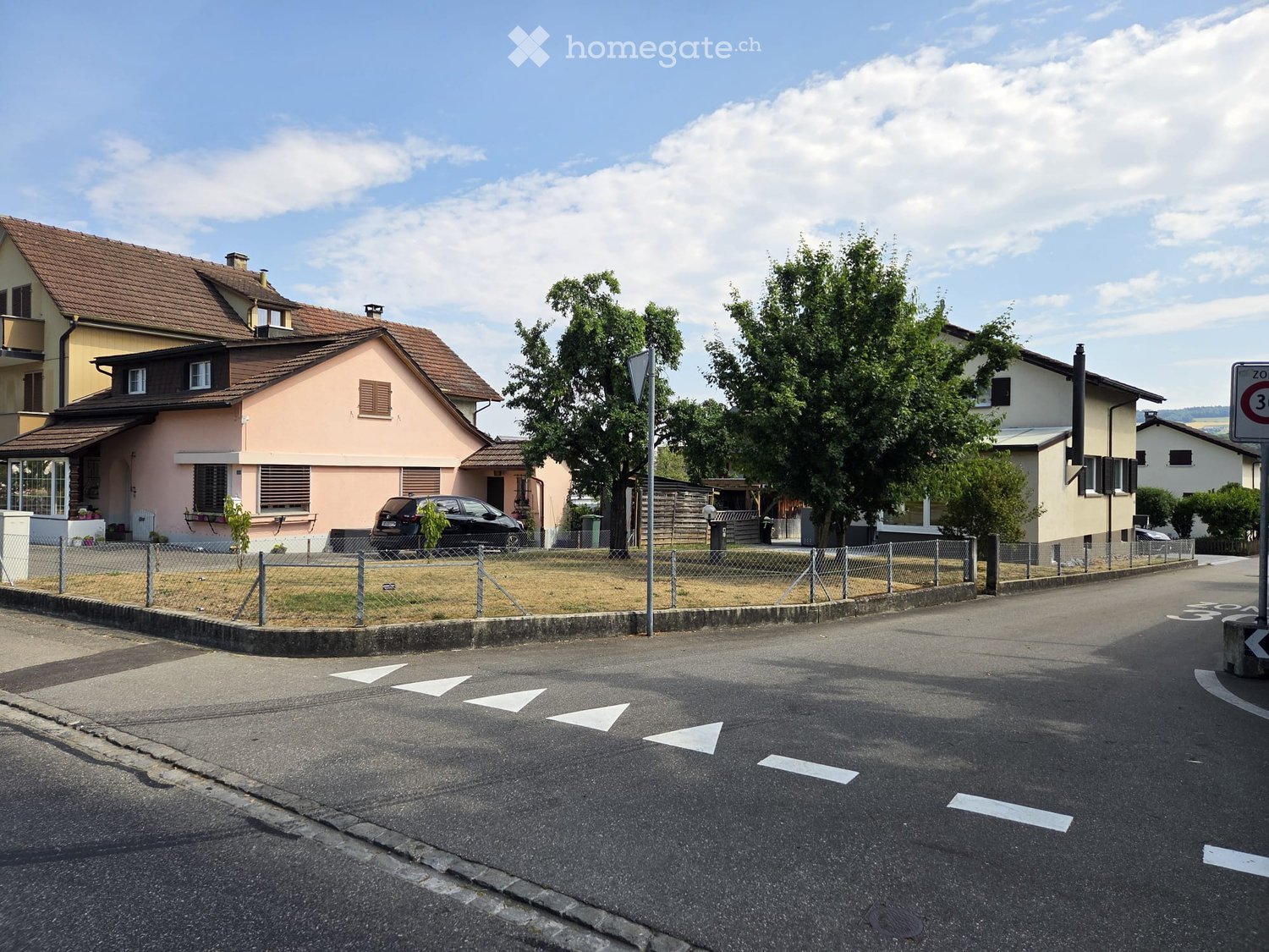 Residential street with multiple houses, including a pink house with a brown roof, a yellow house, and a white house. There is a parked car, a fence, and some greenery.