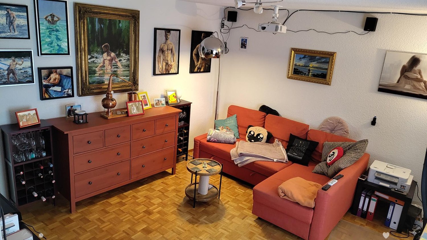 A modern living room with a red sofa, coffee table, wine rack, and various picture frames on the walls. The room features a wooden floor and a projector mounted on the ceiling.