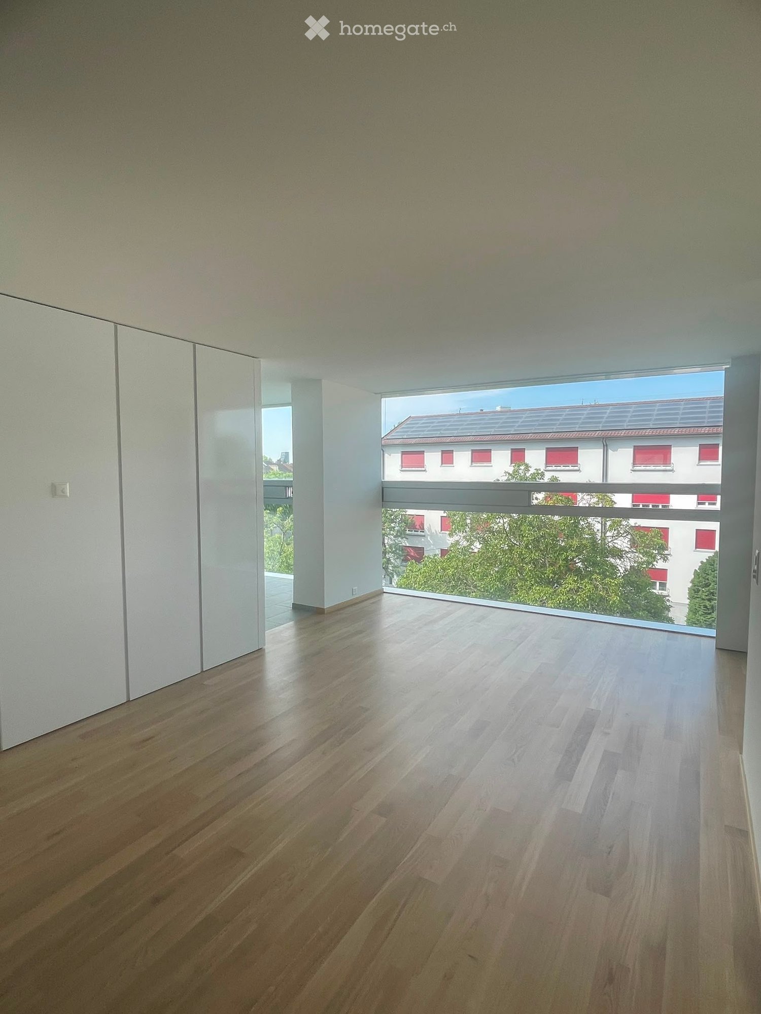 Empty room with light wooden flooring and large windows offering a view of the outside, including trees and buildings.