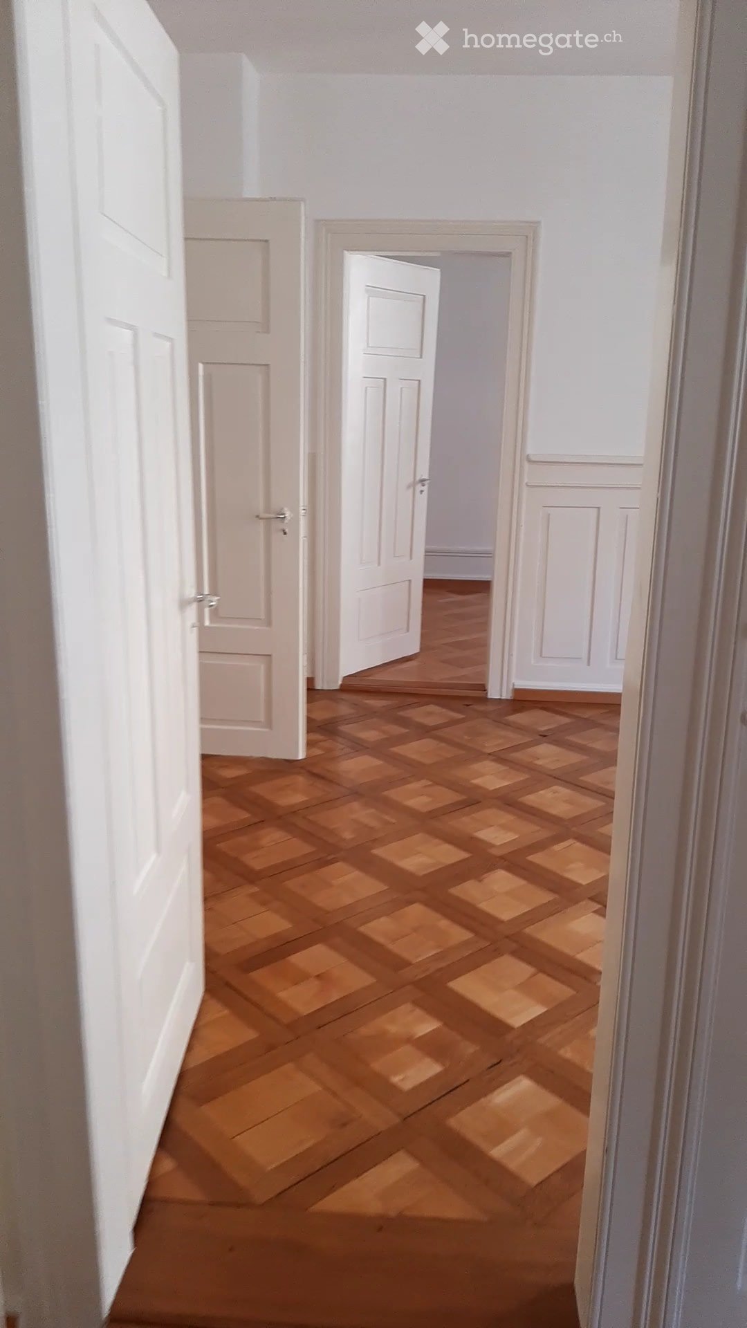 Empty hallway with wooden flooring and white walls, featuring multiple doors and a visible room beyond.