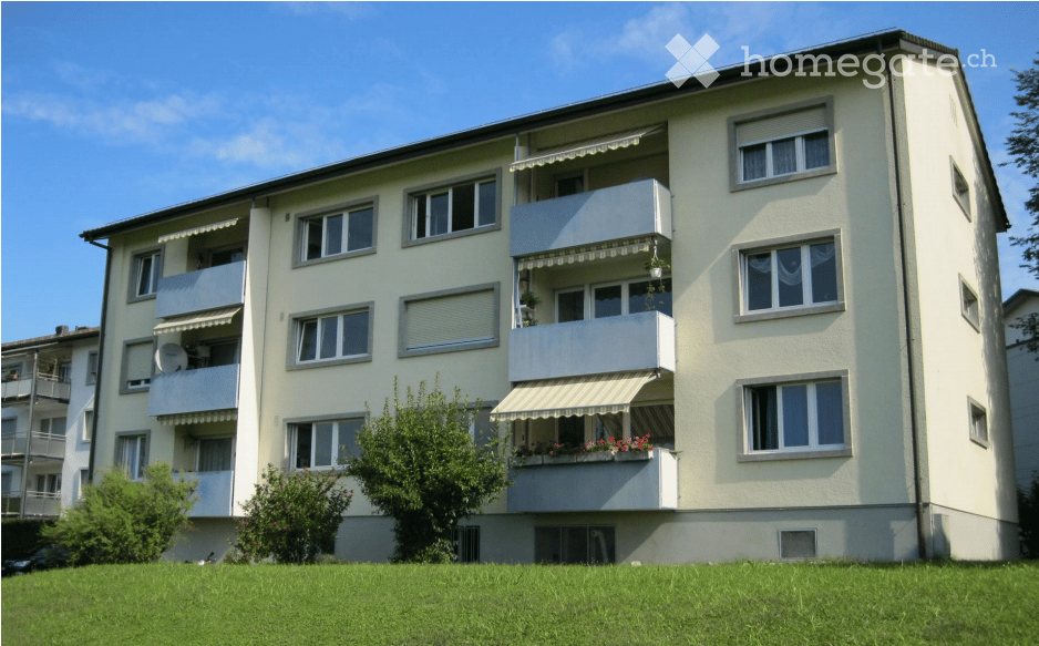 Four-story residential building with multiple balconies, white facade, and green lawn in front.