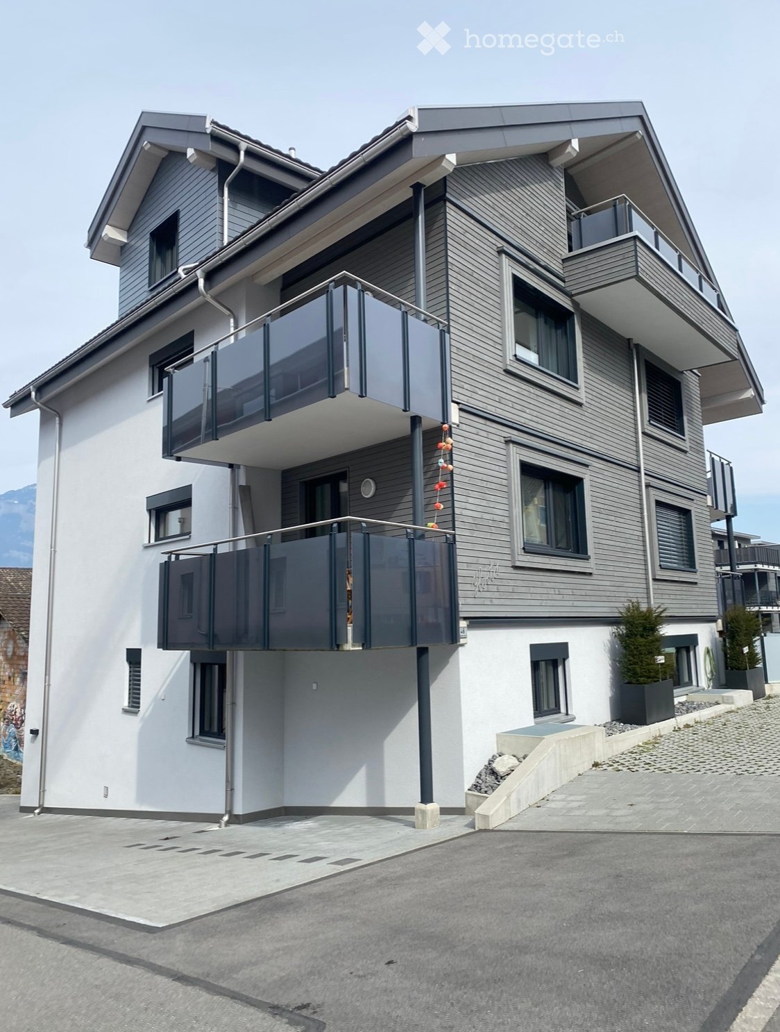 Modern two-story house with grey siding, white lower section, multiple windows, and balconies with glass railings.
