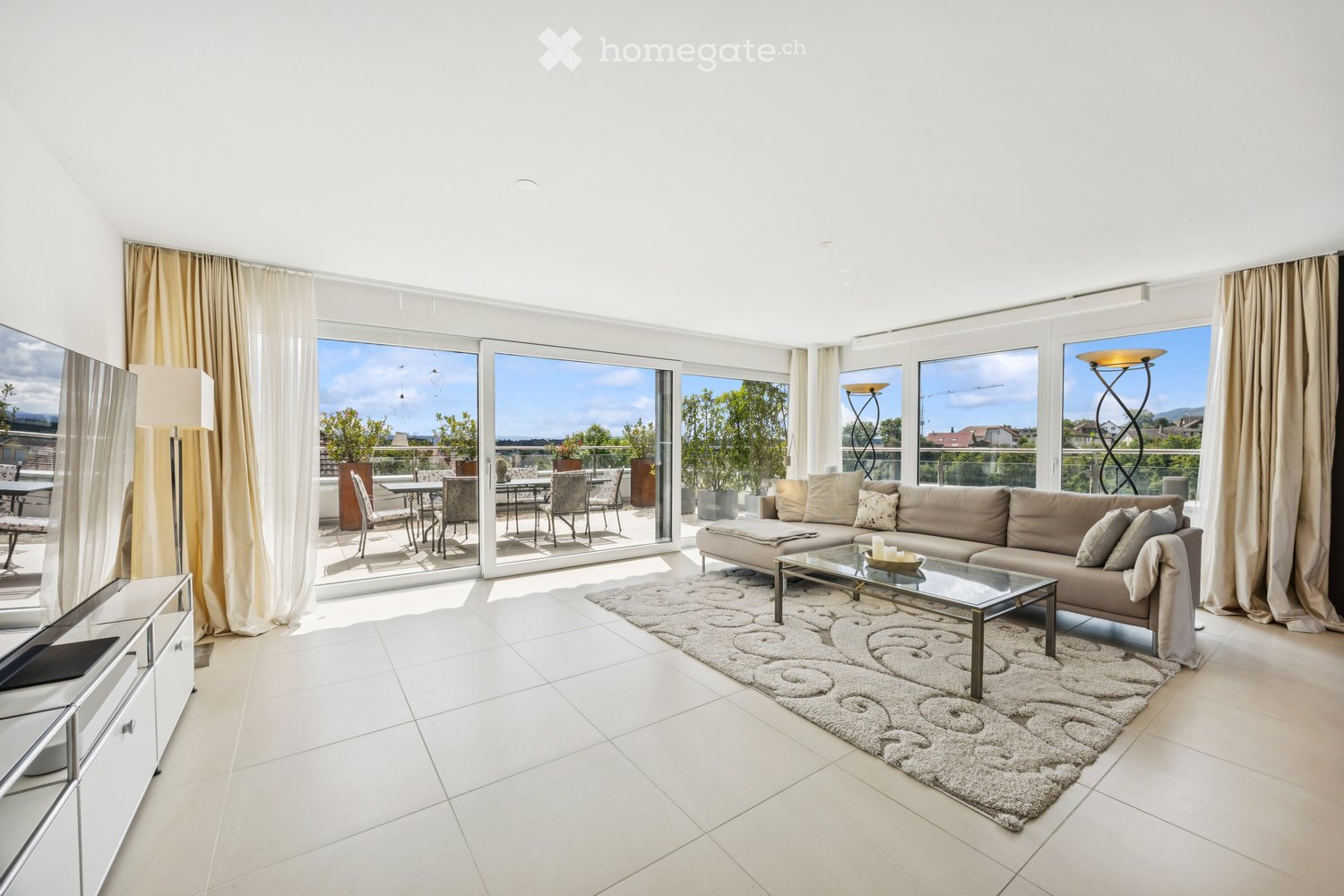 Living Room. Modern living room with large windows and a view, featuring a beige sectional sofa, glass coffee table, floor lamp, TV stand, and access to a balcony with outdoor furniture.